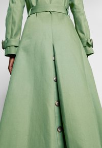 Personne portant un trench-coat vert clair avec des boutons et une ceinture, vue de la taille jusqu'en bas.