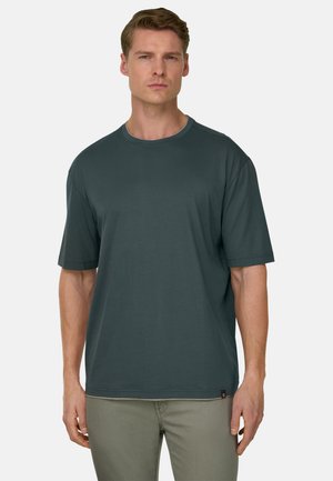 Uomo con capelli corti castano chiaro che indossa una maglietta alla moda oversize a maniche corte di colore verde scuro e pantaloni grigio chiaro, in piedi davanti a uno sfondo semplice.