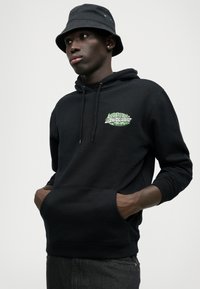 Hanorac negru cu buzunar frontal, având un design grafic verde pe piept. Complementat cu o pălărie tip bucket neagră pentru un look stratificat.