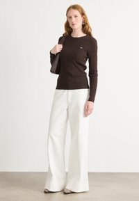 Pull en tricot côtelé marron avec col rond, associé à un pantalon large blanc. Les accessoires comprennent un sac à main et des boucles d'oreilles pendantes.