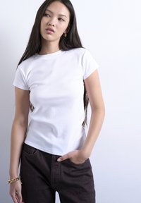 Νέα γυναίκα με μακριά σκούρα μαλλιά, φορώντας λευκό κοντομάνικο μπλουζάκι και μαύρο παντελόνι, στέκεται με το ένα χέρι στην τσέπη μπροστά σε λευκό τοίχο.