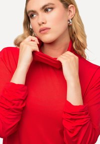 Rode longsleeve top met een hoge kraag. De stof lijkt glad te zijn met geplooide details bij de mouwen, wat zorgt voor een aansluitende silhouet.