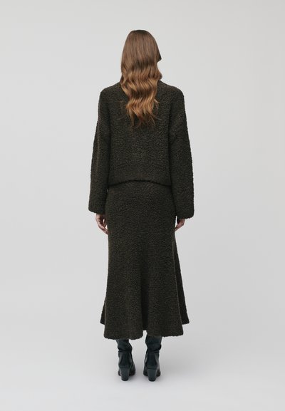 Ensemble en deux pièces comprenant une veste texturée vert olive foncé et une jupe assortie avec un ourlet évasé, accompagnés de bottes à talons noires.