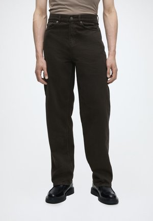 Homme portant un pantalon droit marron foncé taille haute, des chaussures habillées en cuir noir et une chemise beige rentrée.