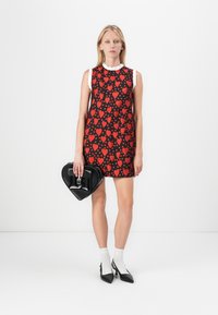 Vestido negro con patrón de corazones rojos, sin mangas, con cuello de volante blanco. El modelo sostiene un bolso negro en forma de corazón, lleva medias blancas y zapatos negros.