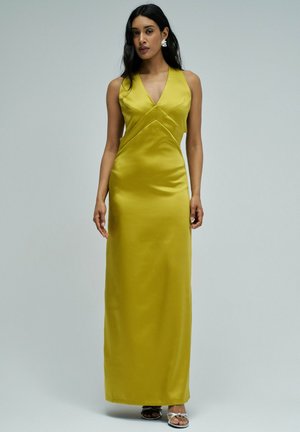 Femme aux longs cheveux foncés portant une robe jaune en satin sans manches avec un col en V et des sandales argentées, debout devant un fond clair uni.