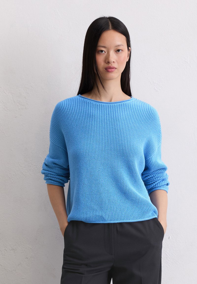 Pull en tricot bleu clair avec une coupe décontractée, texture côtelée, encolure large et manches retroussées, associé à un pantalon sombre.