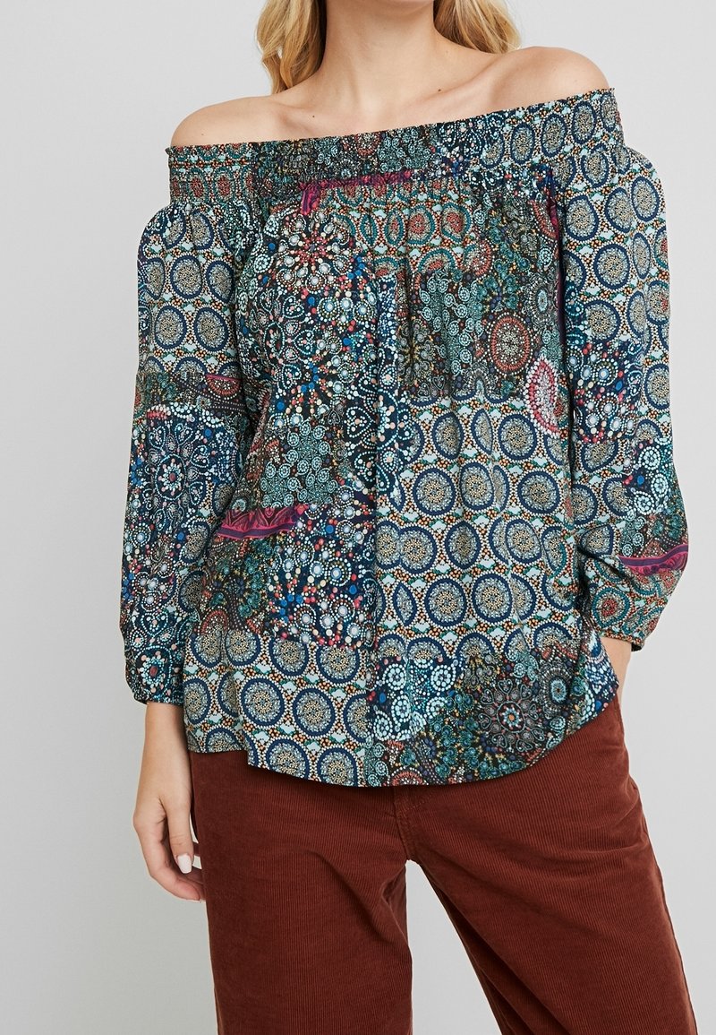 Femme portant un chemisier à épaules dénudées avec des motifs circulaires et floraux multicolores, associé à un pantalon en velours côtelé rouge foncé.