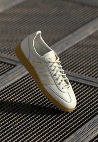 Sneaker in pelle bianca con suola in gomma, tre strisce laterali, logo dorato sulla linguetta e superficie liscia. Design classico a basse top.