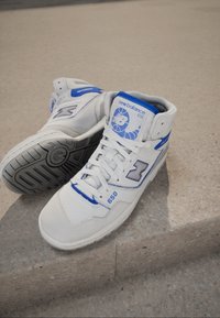 Hvide og blå New Balance 650 høje sneakers med snørebånd, logoer og mønstrede såler, der hviler på en lys betonoverflade.