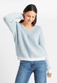 Jasnoniebieski sweter w paski z dekoltem w serek, wykonany z miękkiego materiału, z prążkowaną fakturą i białym wykończeniem, zestawiony z niebieskimi dżinsami.
