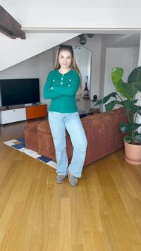 Zeleni rebrasti pulover z zlatimi gumbi, svetlo modri kavbojke in sivi škornji. Ozadje vključuje rjav kavč in lončnico.