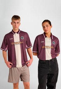 Sportowe koszulki w paski w kolorach bordo i kremowym z krótkimi rękawami. Logo na piersi i kontrastujący kołnierzyk. Model męski w szortach, modelka w czarnych dżinsach.
