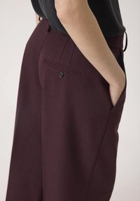 Bordeauxfarbene Hosen aus strukturiertem Stoff, mit einer Seitentasche und einer einzelnen Gesäßtasche mit einer schwarzen Knopfdetails.