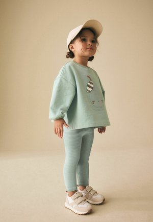 Young child wearing a beige cap, mint green oversized sweater with a pear graphic, striped leggings, and white Velcro sneakers stands against beige background.