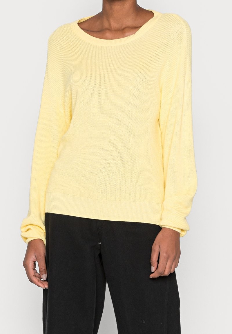 Personne portant un pull en maille ample jaune clair à manches longues et un pantalon noir sur un fond uni.