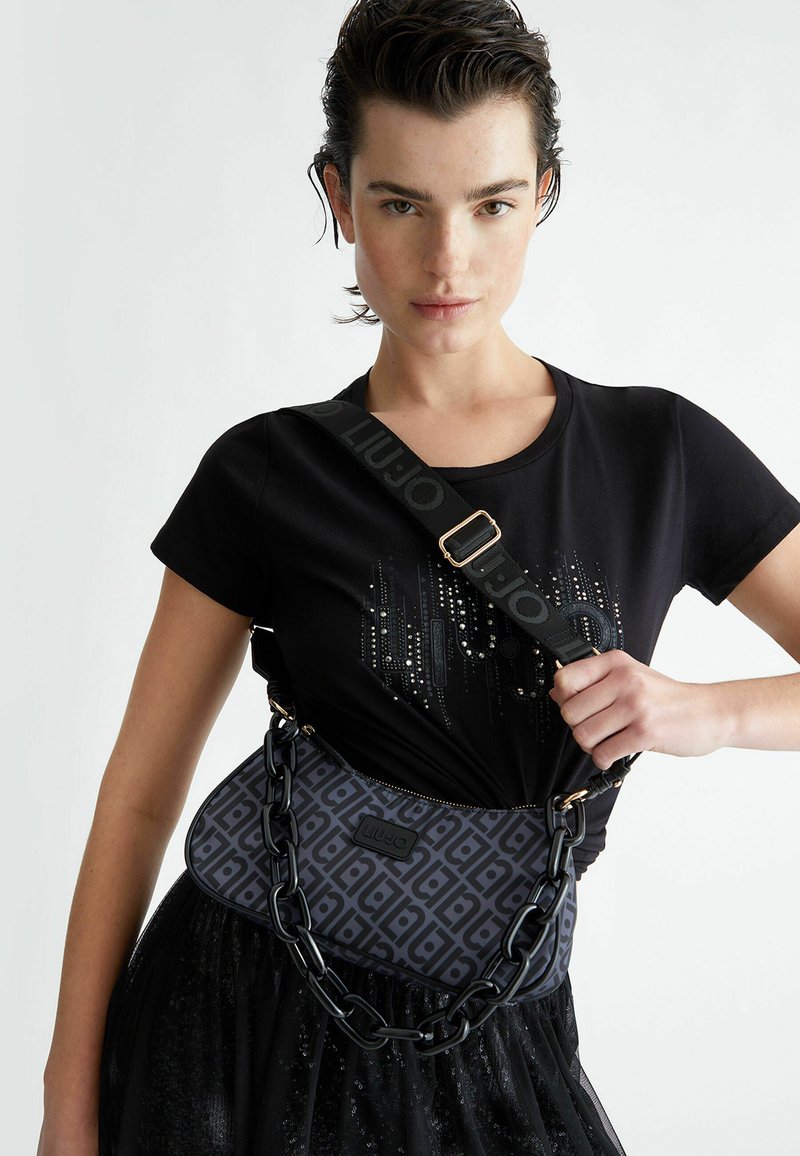 Borsa a spalla nera con motivo geometrico, tessuto texturizzato e dettagli in catena metallica; abbinata a una camicia nera aderente e una gonna di tulle.