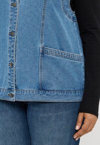 Svetlo modra jeans bunda z gumbnim zapiranjem, dvema stranskima žepoma in strukturirano zasnovo. Nošena preko dolgesleve črne majice z gladko teksturo.