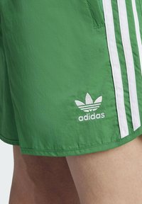 Green athletic shorts made of lightweight material with white vertical stripes and an embroidered white Adidas logo on the left leg.
