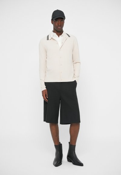 Gai bēšs kardigāns ar pogām, melnbaltajām koledžas strīpām, apvienots ar melniem ceļgala garuma šortiem un elegantiem melniem potīšu zābakiem.