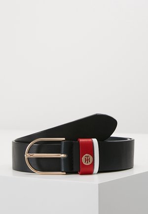 Sort læderbælte med guldfarvet ovalt spænde og røde, hvide og blå læderøjler, hvoraf en har et guld rundt emblem med bogstaver.