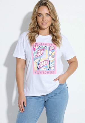Blondynka w białym T-shircie z kolorowym kwiatowym nadrukiem i tekstem "Meadow of Wildflowers,", zestawionym z jasnoniebieskimi dżinsami z brokatem.