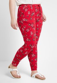 Osoba v červených legínách s kv�ětinovým potiskem a bílých sandálech Birkenstock, stojící s jednou nohou mírně překříženou přes druhou.