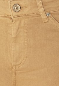 Primer plano de tejido de mezclilla color beige que muestra un detalle de costura en el bolsillo delantero y accesorios de botón de metal. Textura suave con un ligero brillo.