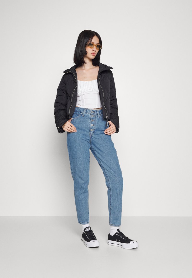 Chaqueta acolchada negra, top corto blanco, jeans de tiro alto de color azul claro con cierre de botones y zapatillas negras con suelas blancas.