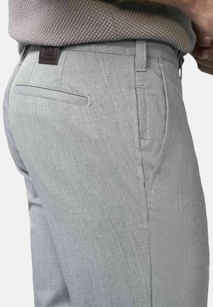 Man draagt lichtgrijze broek met zij- en achterzakken, gecombineerd met een lichtgrijze gebreide trui met structuur, hand rustend dichtbij de taille.