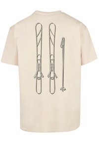 Beige t-shirt featuring a printed design of two ski bindings and a ski pole in gray. The print shows detailed designs and textures.
