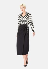 Jupe à enrouler noire nouée à la taille, assortie à un chemisier blanc à grands pois noirs. Tissu lisse et coupe ajustée. Chaussures noires.