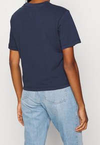 Persona vestida con una camisa de manga corta azul marino y jeans claros, vista desde la espalda contra un fondo liso.