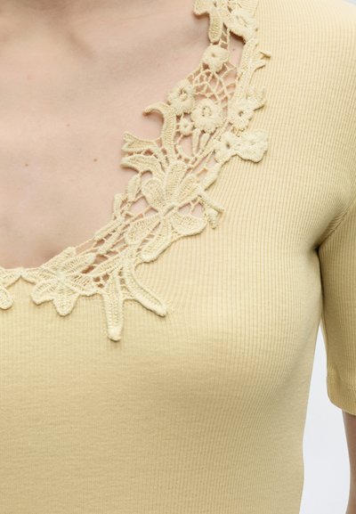 Κοντινό πλάνο μπεζ ριμπ ύφασμα τοπ με λεπτομέρεια από φλοράλ δαντέλα κατά μήκος της V-σχήματος λαιμόκοψης και κοντά μανίκια.