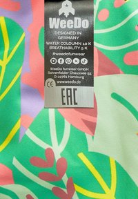 Kleurvol stoffen label met een groen tropisch patroon met roze en gele bladeren. De tekst bevat merkinformatie en verzorgingssymbolen.