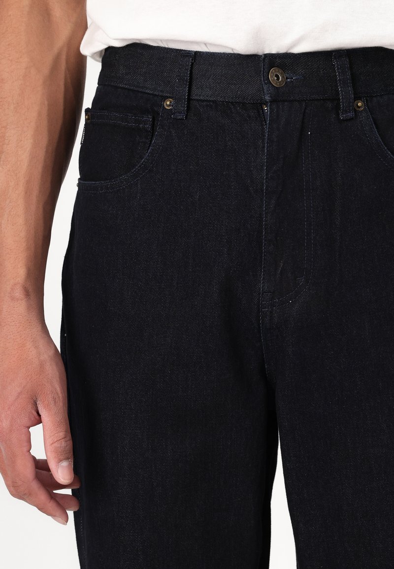 Jeans in denim blu scuro con vestibilità affusolata, dotati di tasche frontali, chiusura con bottone metallico e cuciture a vista. Texture liscia.
