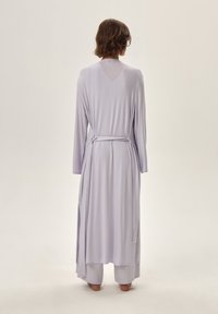 Długa lawendowa szata z długimi rękawami, z paskiem w talii i luźnym krojem, wykonana z miękkiej, zwiewnej tkaniny. Widoczne boczne rozcięcia u dołu.