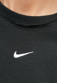 Μαύρο μπλουζάκι με στρογγυλή λαιμόκοψη και ξεφτισμένες άκρες, με λευκό κεντημένο λογότυπο Nike στο στήθος. Η υφή είναι λεία και ελαστική.