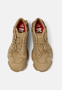 Sneakers scamosciati color cammello con un design arrotondato, superficie testurizzata e lacci prominenti. Gli interni presentano accenti rossi con branding.