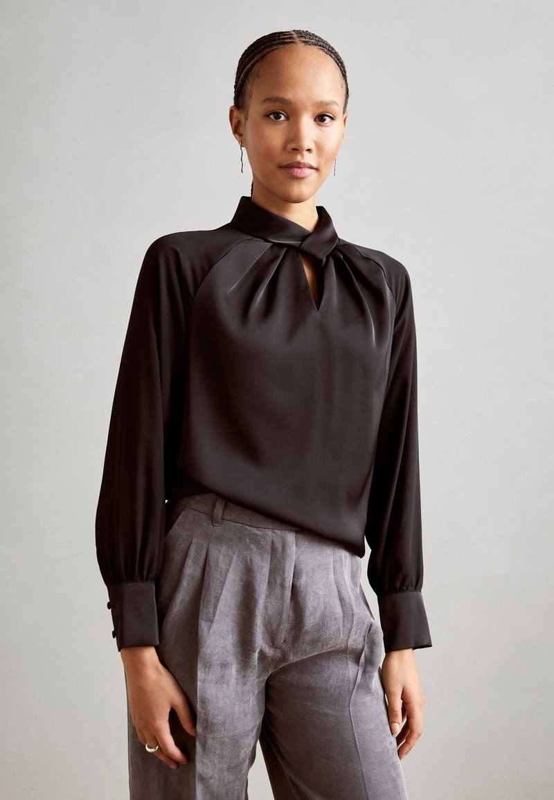 Schwarze Bluse mit langen Ärmeln, hohem Kragen und einem Schlüsselloch-Ausschnitt. Der Stoff hat eine glatte Textur. Kombiniert mit hellgrauen, plissierten Hosen.