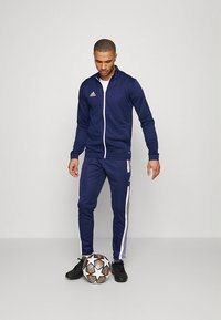 Νavy φόρμα με λευκές ρίγες, κατασκευασμένη από ελαφρύ ύφασμα. Το μοντέλο στέκεται με μια ποδοσφαιρική μπάλα, φορόντας μαύρα αθλητικά παπούτσια.