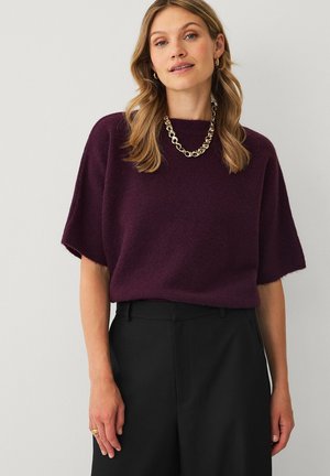 Kvinde med skulderlangt blondt hår iført en kortærmet mørkelilla sweater, sorte bukser, guldkæde halskæde og guldringe øreringe.