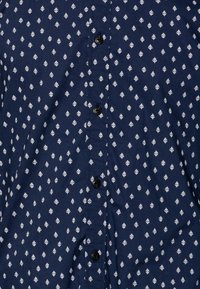 Camisa de botones azul marino con un patrón de rombos blancos. Fabricada con una tela de superficie ligeramente texturizada y seis botones negros en el frente.