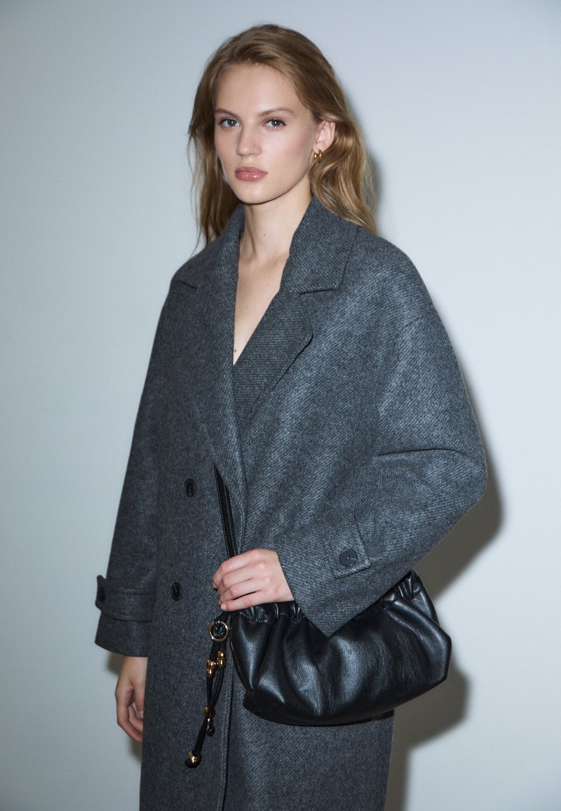 Cappotto grigio in lana con ampi revers e bottoni, abbinato a una borsa a mano nera in pelle con design arricciato e dettagli in oro.