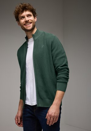 Veste zippée verte texturée avec des poignets et un ourlet côtelés, portée sur un t-shirt blanc, associée à un jean bleu foncé.