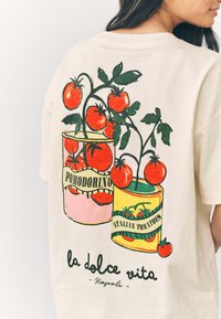 Krém színű póló, melyen egy illusztrált grafika látható két paradicsomos dobozzal, piros paradicsomokkal és zöld levelekkel, "Pomodorino" és "Olasz paradicsomok" feliratokkal.