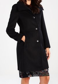 Svart ullrock med stor krage, knappframsida och sidofickor. Den eleganta designen kompletterar en mönstrad kjol. Rena linjer och subtil textur.