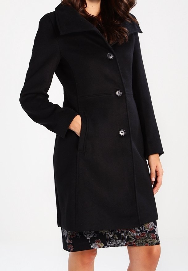 Svart ullrock med stor krage, knappframsida och sidofickor. Den eleganta designen kompletterar en mönstrad kjol. Rena linjer och subtil textur.