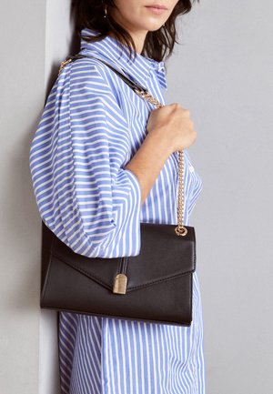 Woman in blue and white striped shirt holding a black leather purse with a gold chain strap over shoulder against gray wall.