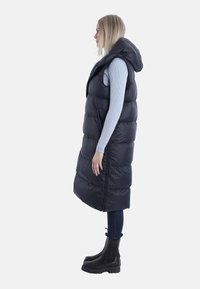 Schwarze Steppweste mit Kapuze, ausgestattet mit einem langen, gesteppte Design und Reißverschluss. Getragen über einem hellblauen Langarmoberteil und dunklen Jeans.